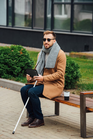 Homem sentado no banco da praça com sua BrailleOne ao lado sua bengala